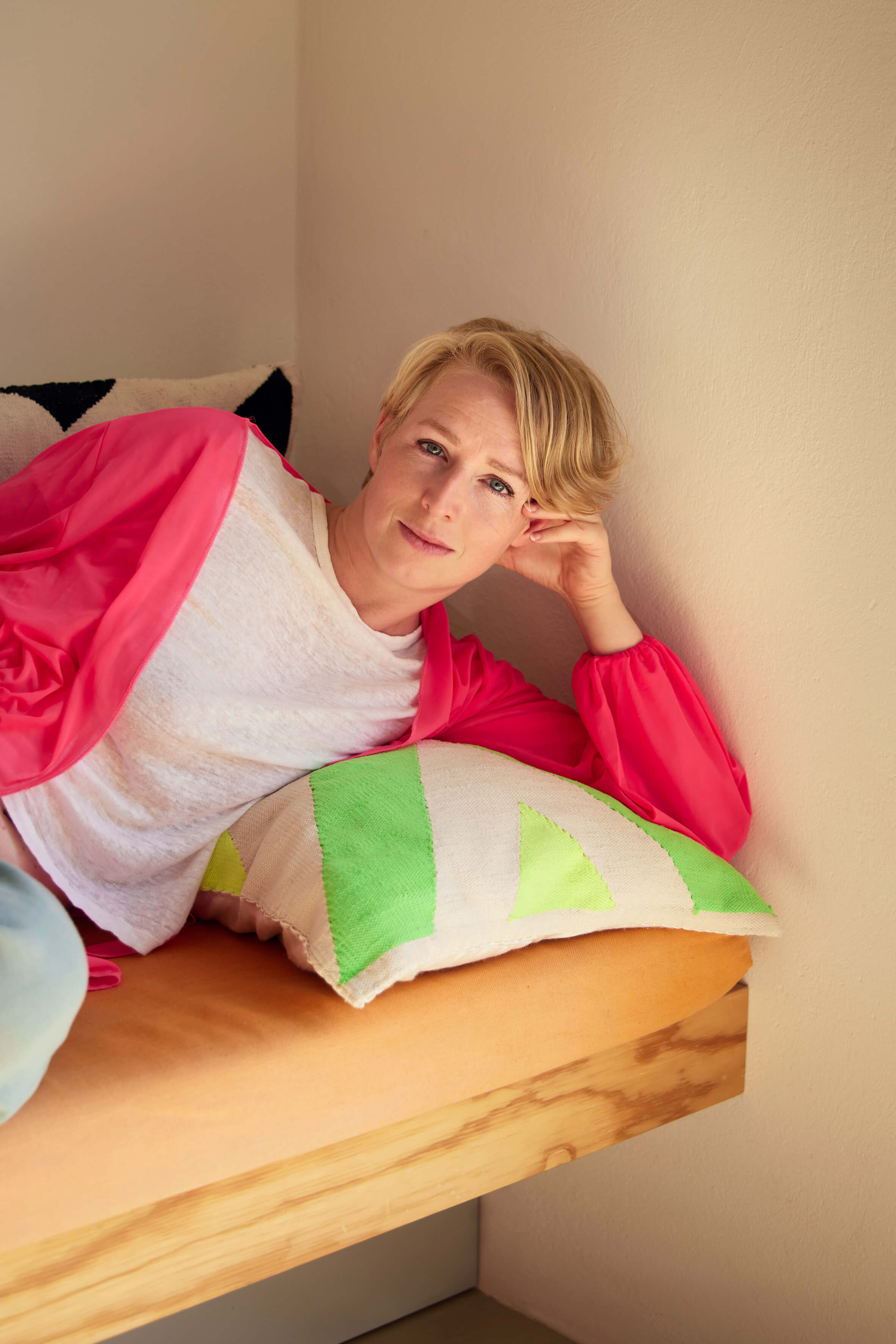 Eine Person mit kurzen blonden Haaren, weißem T-Shirt und langem rosa Cardigan sowie hellblauen Jeans sitzt auf einem Sofa mit bunten Kissen und lächelt in die Kamera.
