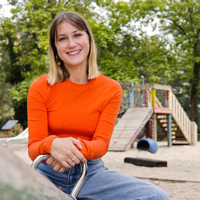 Eine Person mit schulterlangen blonden Haaren, orangenem Pullover und blauer Jeans sitzt auf einer Wippe an einem Kinderspielplatz und lächelt in die Kamera. 
