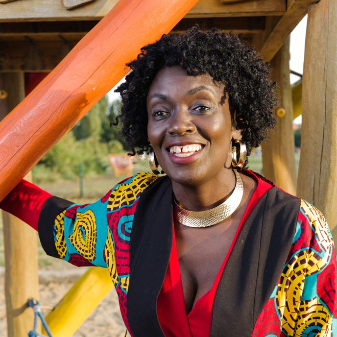 Eine Person mit kurzen, dunklen Rastas und einem bunten Kleid steht an einem Bauturm an einem Kinderspielpatz und lächelt in die Kamera. 