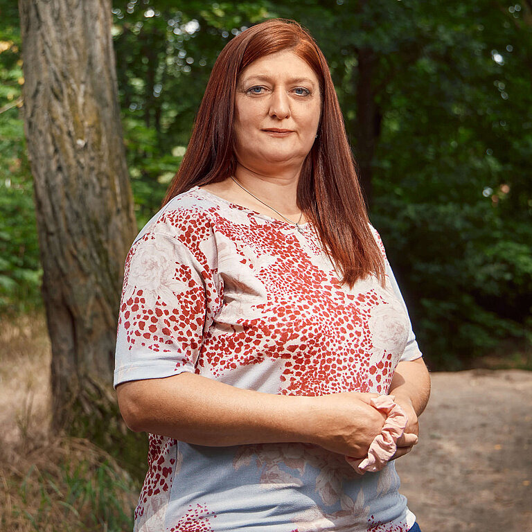 Eine Person mit langen roten Haaren, hellblau-rot gemusterten Shirt und dunklen Jeans steht vor einem Waldweg und lächelt in die Kamera.