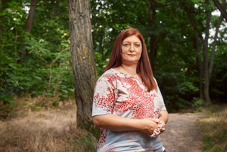Eine Person mit langen roten Haaren, hellblau-rot gemusterten Shirt und dunklen Jeans steht vor einem Waldweg und lacht in die Kamera.