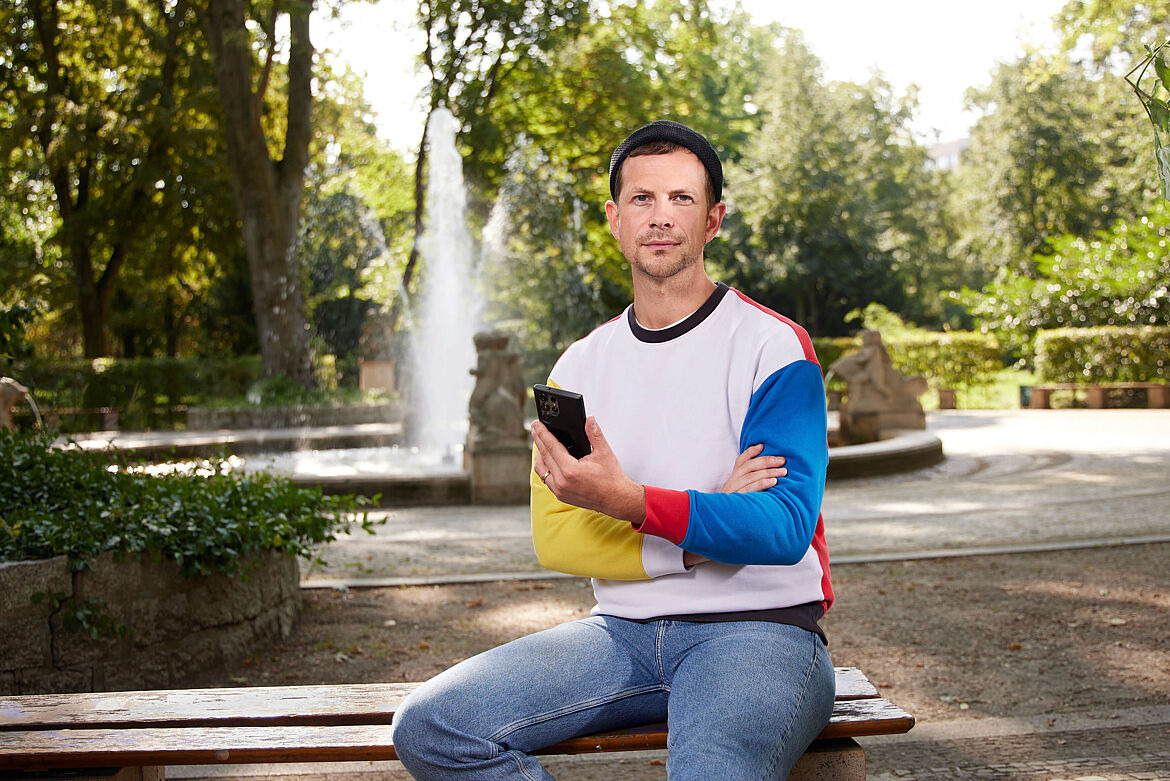 Eine Person mit kurzen dunklen Haaren, dunkelblauer Mütze und bunt gemusterten Longsleeve sitzt mit einem Handy in der Hand an einem Stadtbrunnen und schaut in die Kamera. 
