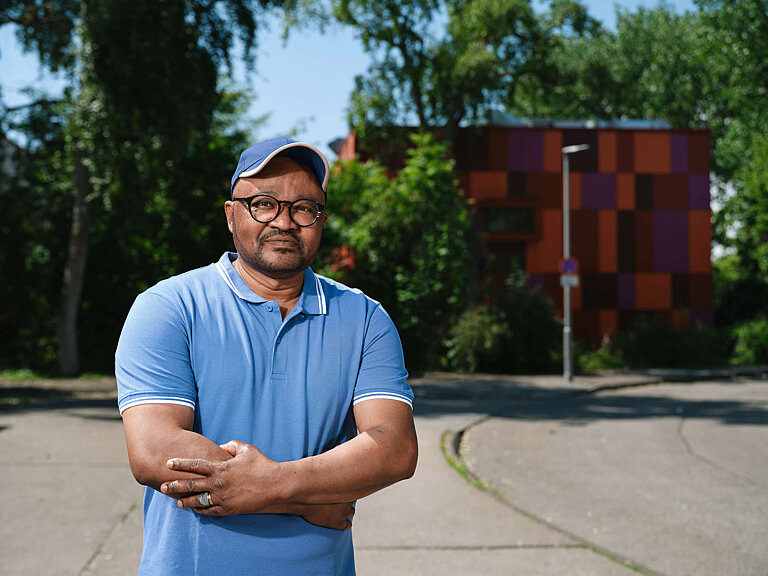 Eine Person mit schwarzer Brille, blauen Cap und blauen Polo-Shirt steht vauf einer Straße. Hinter ihm sieht man grüne Bäume und ein braunes Haus.