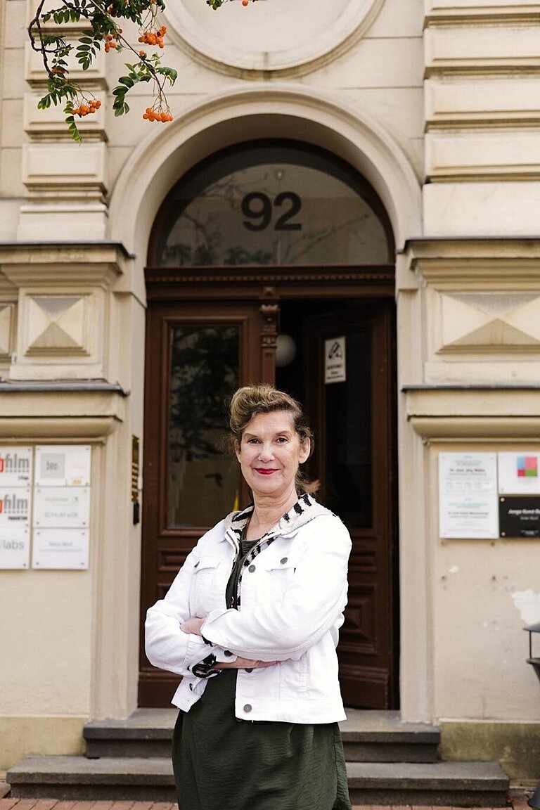 Eine Person mit dunklem Pferdeschwanz, schwarzem Kleid und weißer Jeansjacke steht mit verschränkten Armen vor einem Eingang eines Altbaus und lächelt in die Kamera. 