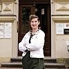 Eine Person mit dunklem Pferdeschwanz, schwarzem Kleid und weißer Jeansjacke steht mit verschränkten Armen vor einem Eingang eines Altbaus und lächelt in die Kamera. 