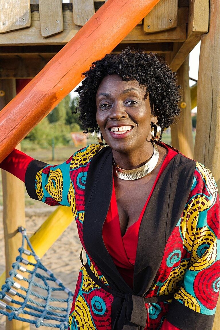 Eine Person mit kurzen, dunklen Rastas und einem bunten Kleid steht an einem Bauturm an einem Kinderspielpatz und lächelt in die Kamera. 