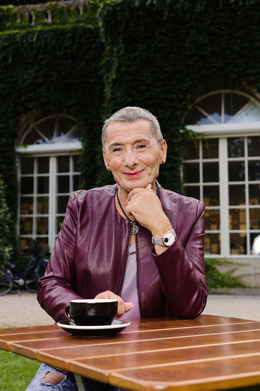 Eine Person mit ganz kurzen grauen Haaren und violetter Lederjacke sitzt mit einem Kaffee am Gartentisch und lächelt in die Kamera. 