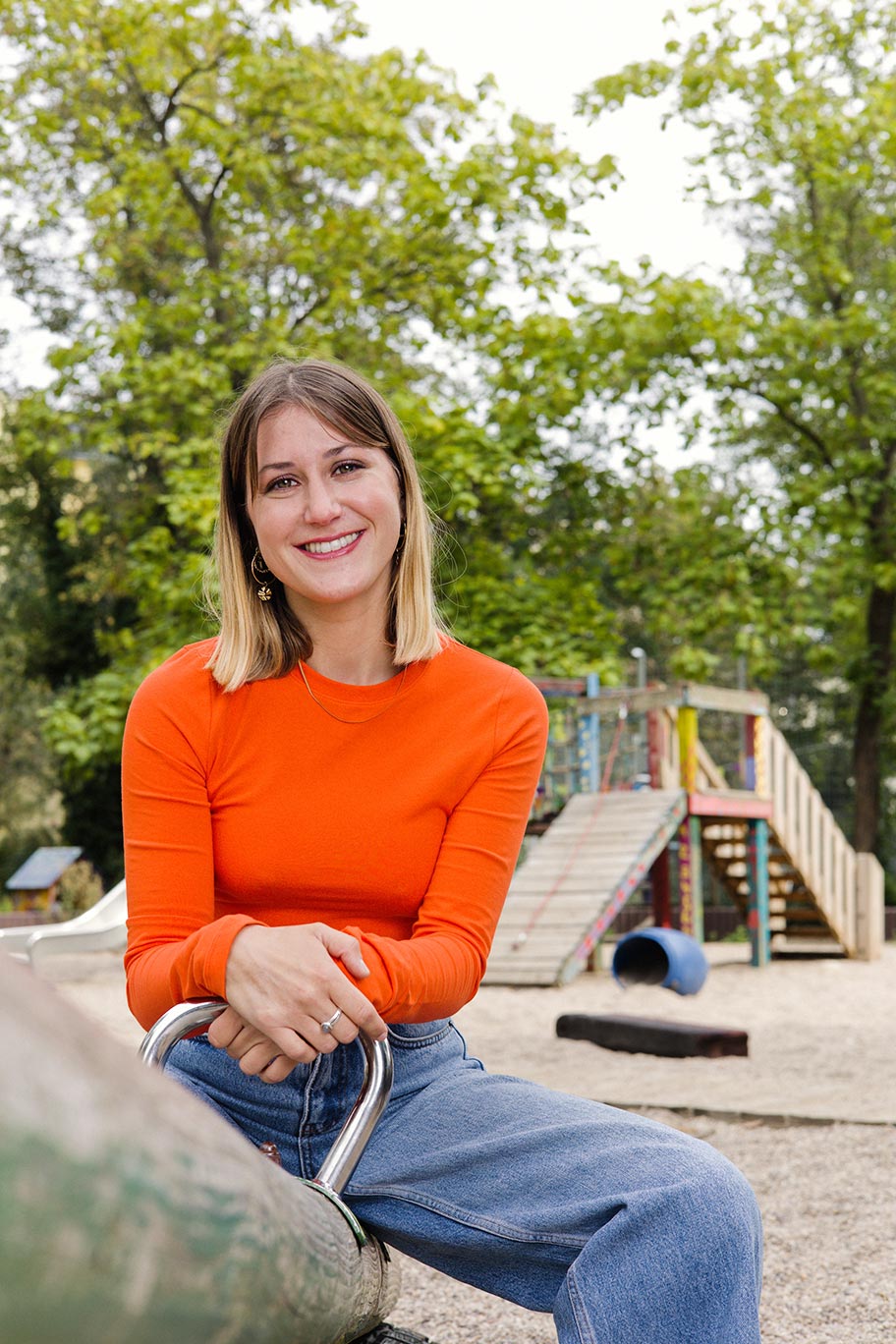 Eine Person mit schulterlangen blonden Haaren, orangenem Pullover und blauer Jeans sitzt auf einer Wippe an einem Kinderspielplatz und lächelt in die Kamera. 