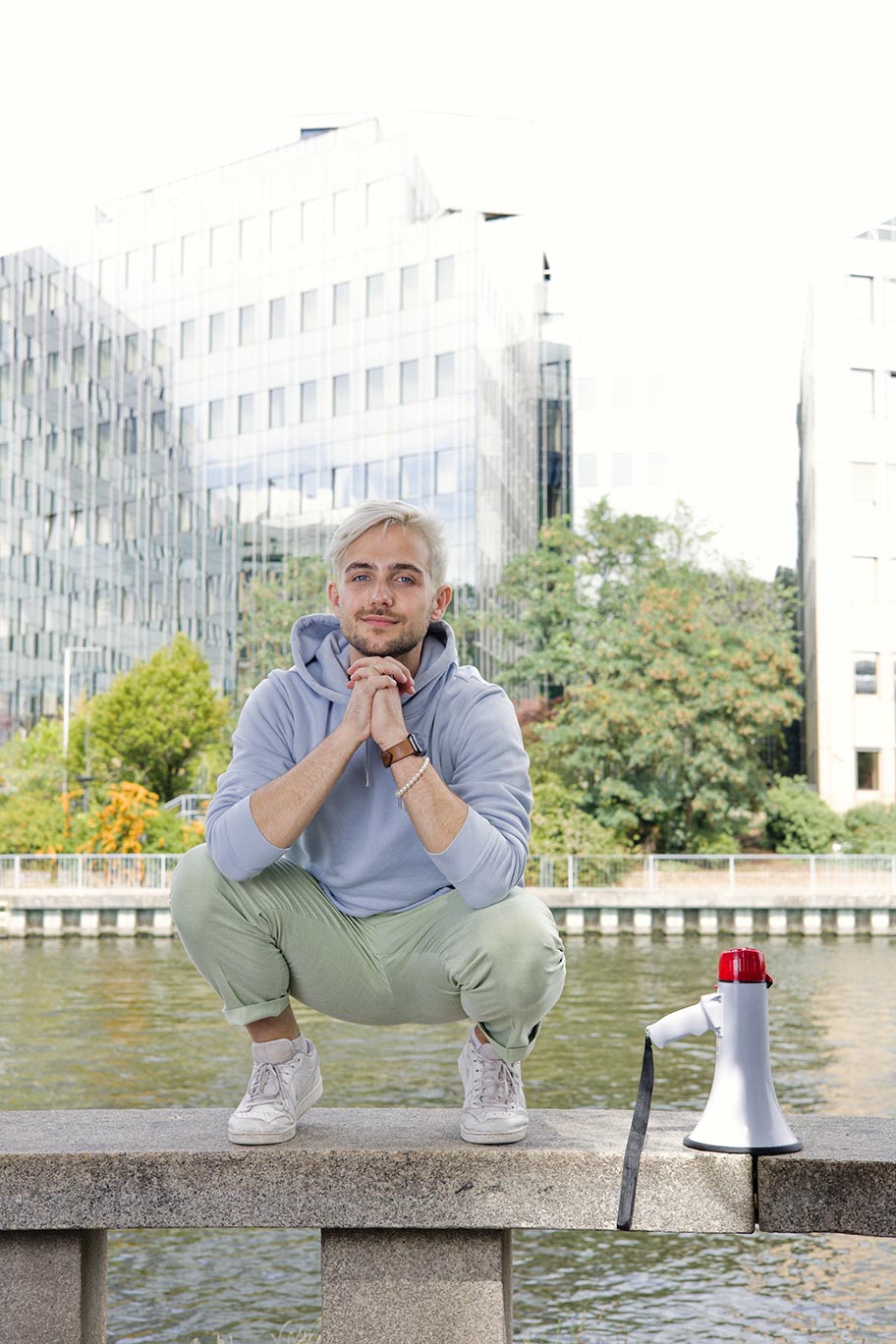 Eine Person mit blonden Haaren und dunklen Bart, hellblauen Hoodie sowie grüner Hose sitzt vor einem Glasgebäude und lächelt in die Kamera. 